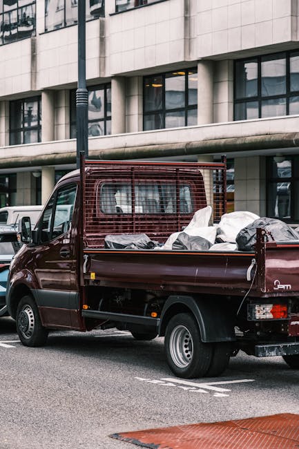 A waste collection truck with a primarily white body and metallic accents is parked on a narrow cobblestone street, positioned with its rear lift open and ready to load waste. The truck's surface show
