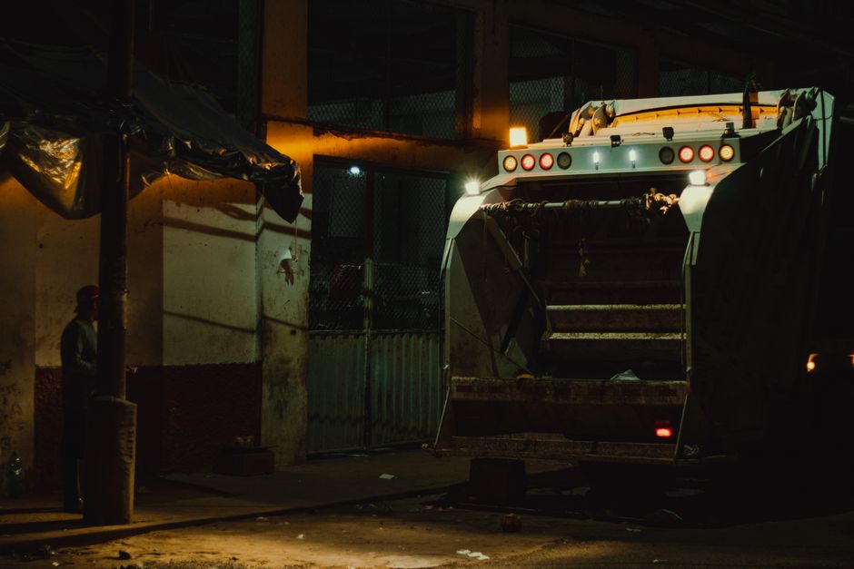 A waste collection worker dressed in high-visibility orange workwear with reflective stripes is seen from above, pushing a small, wheeled waste bin along a paved street. The bin, positioned in the mid