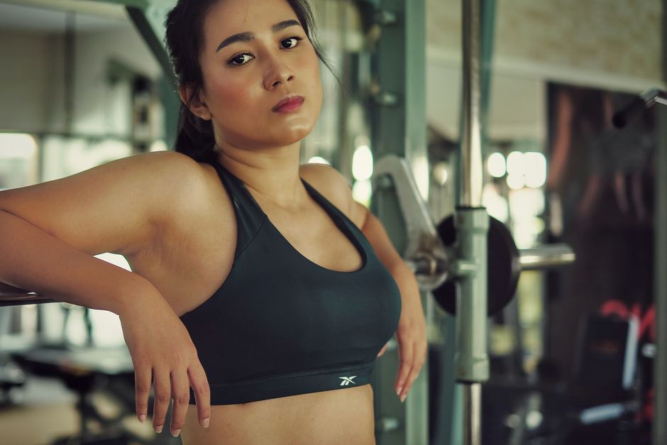 A young woman with medium skin tone and dark hair tied back, wearing a black sports bra with a subtle logo on the front, stands in a modern gym environment. She is leaning slightly on her left arm, wh